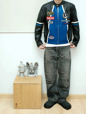 Person wearing a black and blue leather jacket with various logos, standing next to a wooden box with decorative items.