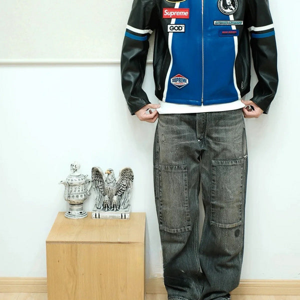 Person wearing a black and blue leather jacket with various logos, standing next to a wooden box with decorative items.