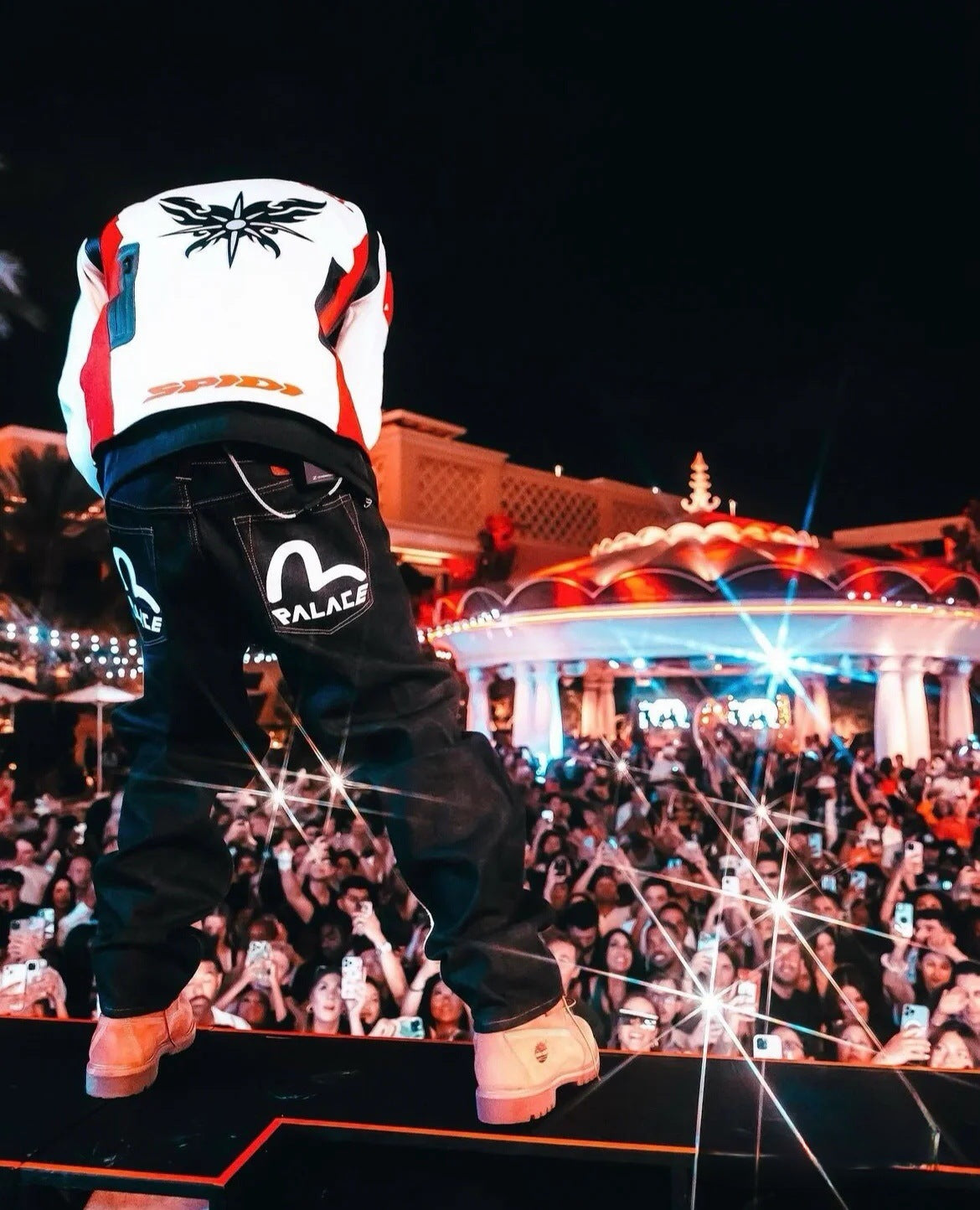 Person wearing a white helmet with a logo and black pants with a brand logo, standing on a stage in front of an audience at night.