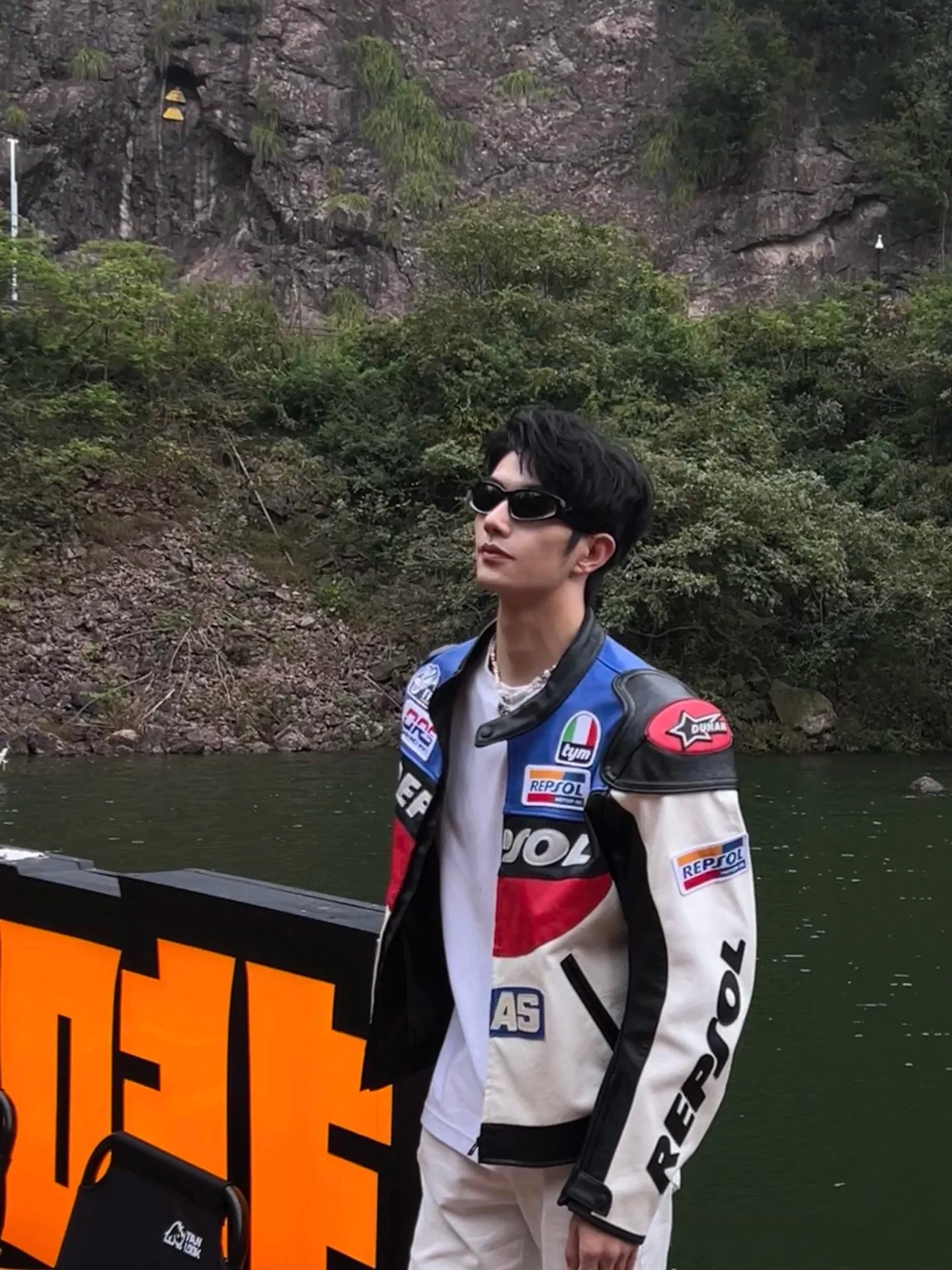 Person wearing a colorful jacket with sponsor logos near a body of water and rocky cliff.